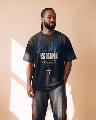 Jesus Is King faded black tee, front print, man with braided hair looking to the side, hands relaxed in warm window light