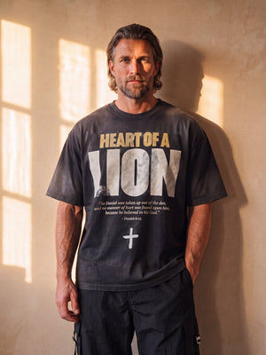 Heart of a Lion faded black tee, front print, man with a beard and one hand in his pocket against a sunlit wall