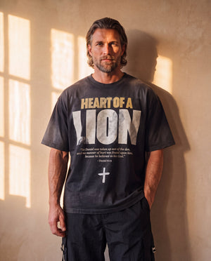 Heart of a Lion faded black tee, front print, man with a beard and one hand in his pocket against a sunlit wall