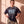 Pray Until Something Happens faded black tee, front print, woman with a hair bun pulling the hem down with both hands over camo pants