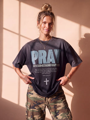 Pray Until Something Happens faded black tee, front print, woman with a hair bun pulling the hem down with both hands over camo pants