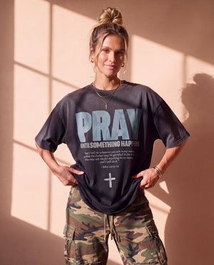 Pray Until Something Happens faded black tee, front print, woman with a hair bun pulling the hem down with both hands over camo pants
