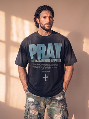 Pray Until Something Happens faded black tee, front print, man with curly hair and hands in his pockets, styled with ripped camo cargo pants