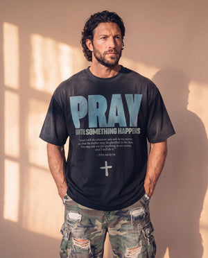 Pray Until Something Happens faded black tee, front print, man with curly hair and hands in his pockets, styled with ripped camo cargo pants