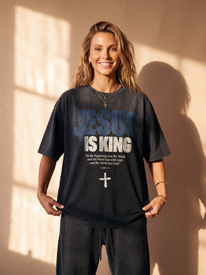 Jesus Is King faded black tee, front print, smiling woman with long wavy hair holding the hem, John 1:1 verse and cross in view