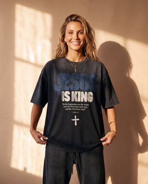 Jesus Is King faded black tee, front print, smiling woman with long wavy hair holding the hem, John 1:1 verse and cross in view