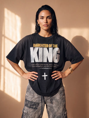 Daughter of the King faded black tee, front print, woman with wet look hair and both hands on her hips, worn over washed cargo pants