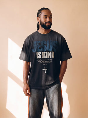 Jesus Is King faded black tee, front print, man with braided hair looking to the side, hands relaxed in warm window light