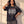 In the Beginning God faded black tee, front print, worn oversized by a woman with one hand in her hair and one on her hip in window light