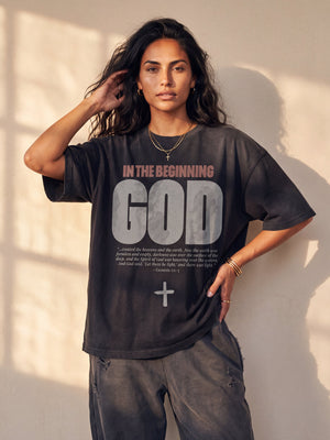 In the Beginning God faded black tee, front print, worn oversized by a woman with one hand in her hair and one on her hip in window light