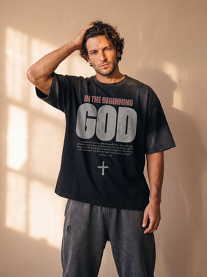 In the Beginning God faded black tee, front print, worn by a man running one hand through his hair against a sunlit wall