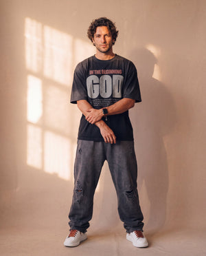 In the Beginning God faded black tee, full body view of a man with crossed wrists in ripped baggy jeans and sneakers, Christian streetwear