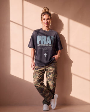 Pray Until Something Happens faded black tee, full body view of a woman leaning on a wall in camo pants and sneakers, Christian streetwear