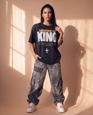 Daughter of the King faded black tee, full body view of a woman in washed cargo pants and sneakers, one hand in her pocket by a sunlit wall