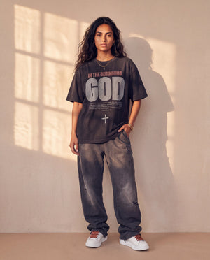 In the Beginning God faded black tee, full body view of a woman in baggy faded jeans and sneakers, one hand in her pocket by a sunlit wall