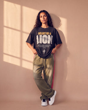 Heart of a Lion faded black tee, full body view of a woman leaning on a sunlit wall in striped track pants and sneakers