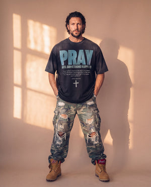Pray Until Something Happens faded black tee, full body view of a man in ripped camo cargo pants and work boots, hands in his pockets