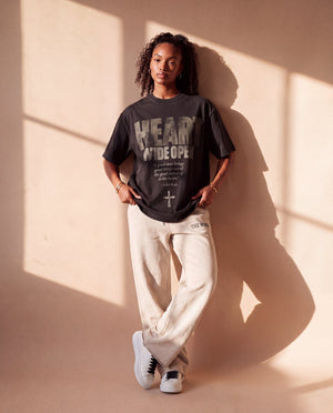 Heart Wide Open faded black tee, full body view of a woman leaning on a sunlit wall, legs crossed, in sweatpants and sneakers