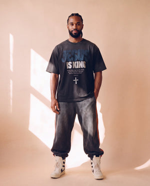 Jesus Is King faded black tee, full body view of a man with braids in sweatpants and high top sneakers, hands in his pockets