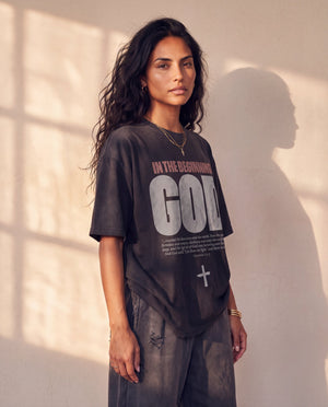 In the Beginning God faded black tee, three quarter angle on a woman with long curly hair, showing the boxy fit and the Genesis verse