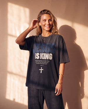 Jesus Is King faded black tee, smiling woman tucking her hair behind her ear, oversized fit with dropped shoulders