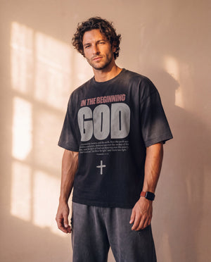 In the Beginning God faded black tee, front print, man standing relaxed with a smartwatch, Christian streetwear with a boxy oversized fit