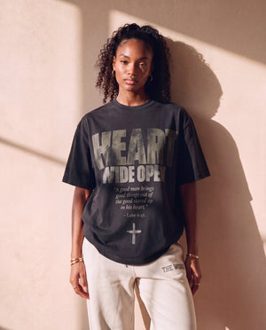 Heart Wide Open faded black tee, front print, woman standing relaxed against a sunlit wall, boxy fit over THE WORD sweatpants