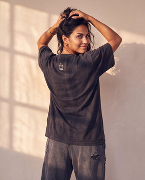 In the Beginning God faded black tee, back view of a woman with both hands in her hair, small crown logo below the collar