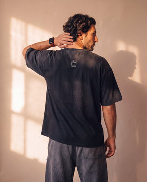 In the Beginning God faded black tee, back view of a man with a hand on his neck, showing the boxy cut and small crown logo