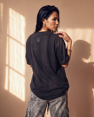 Daughter of the King faded black tee, back view of a woman with a hand on her shoulder, small crown logo below the collar
