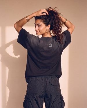 King of Kings faded black tee, back view of a woman with both hands in her hair, looking over her shoulder, crown logo below the collar