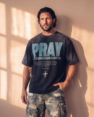 Pray Until Something Happens faded black tee, front print, man leaning on a sunlit wall with one hand in his pocket and his gaze to the side