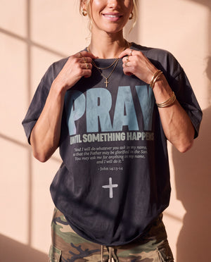 Pray Until Something Happens faded black tee, front print, smiling woman holding the collar, cross necklace above the PRAY lettering