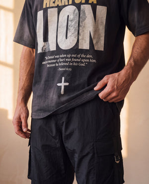 Heart of a Lion faded black tee, close crop on a man holding the hem, the Daniel 6:23 verse and cross above his cargo pants