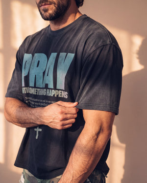 Pray Until Something Happens faded black tee, close up on a man's arm pinching the sleeve, PRAY UNTIL SOMETHING HAPPENS lettering beside it