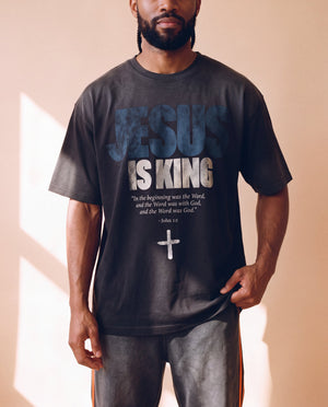 Jesus Is King faded black tee, front print close crop on a man holding the hem, John 1:1 verse and cross, Christian streetwear