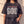 In the Beginning God faded black tee, front print in full on a woman, Genesis 1:1–5 verse and cross, one hand tugging the hem