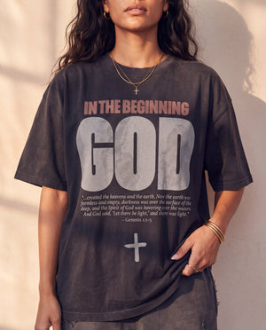 In the Beginning God faded black tee, front print in full on a woman, Genesis 1:1–5 verse and cross, one hand tugging the hem
