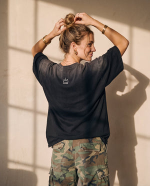 Pray Until Something Happens faded black tee, back view of a woman with both hands in her hair bun, small crown logo below the collar
