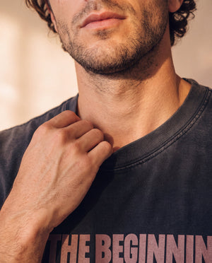 In the Beginning God faded black tee, close up of a man's jaw and hand at the ribbed collar, the top of the print just in frame