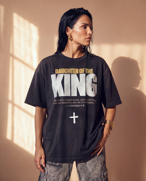 Daughter of the King faded black tee, front print, woman looking to the side by a sunlit wall, the oversized fit hanging loose
