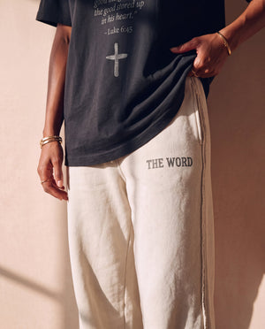 Heart Wide Open faded black tee, close crop on a woman lifting the hem over THE WORD sweatpants, the verse and cross above