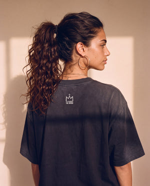 King of Kings faded black tee, back view of a woman with a curly ponytail and hoop earrings, small crown logo under the neckline