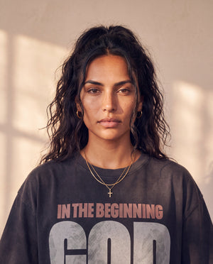 In the Beginning God faded black tee, close up portrait of a woman with curly hair and a cross necklace above the large GOD lettering