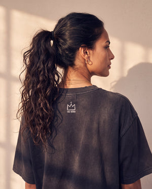 In the Beginning God faded black tee, back view of a woman with a curly ponytail, crown logo and THE WORD under the neckline