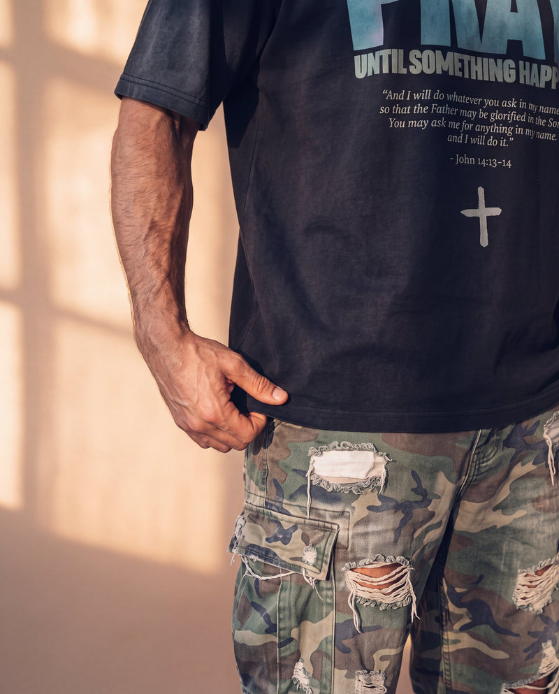 Pray Until Something Happens faded black tee, close up of the hem on a man's hip, the verse and cross just above his hand and camo pants
