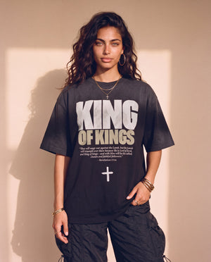 King of Kings faded black tee, front print, woman standing with a hand in her pocket, Revelation 17:14 verse and cross, Christian streetwear