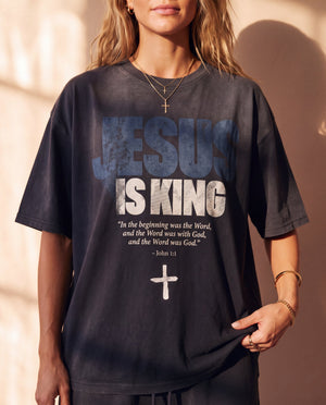 Jesus Is King faded black tee, front print in full on a woman with a cross necklace, John 1:1 verse and cross, Christian streetwear