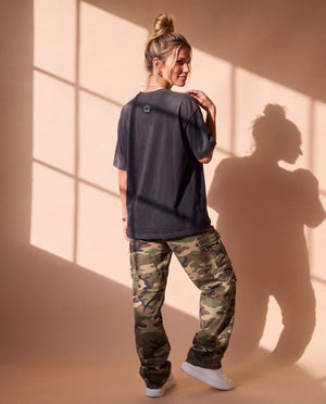 Pray Until Something Happens faded black tee, back view of a woman in camo pants looking over her shoulder, crown logo at the collar