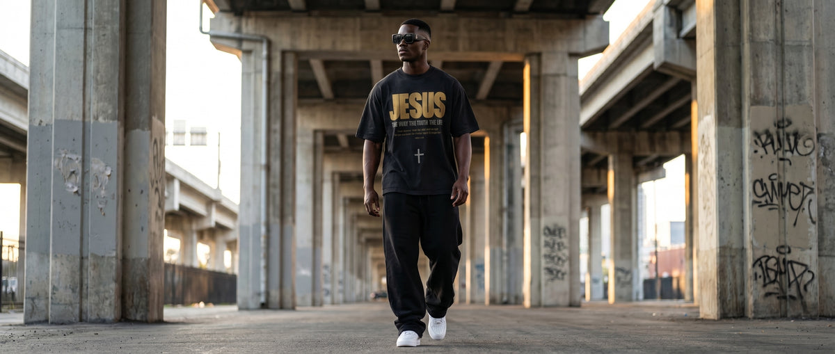 Man in a black and gold JESUS tee walking beneath a concrete overpass